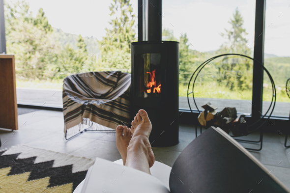 Beautiful female feet and book on background of warm fireplace and big window in modern cozy chalet