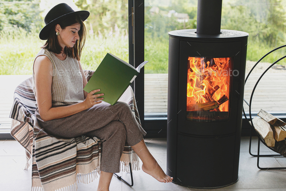 Beautiful stylish woman reading book on chair at warm burning fireplace, cozy vacation. Leisure time