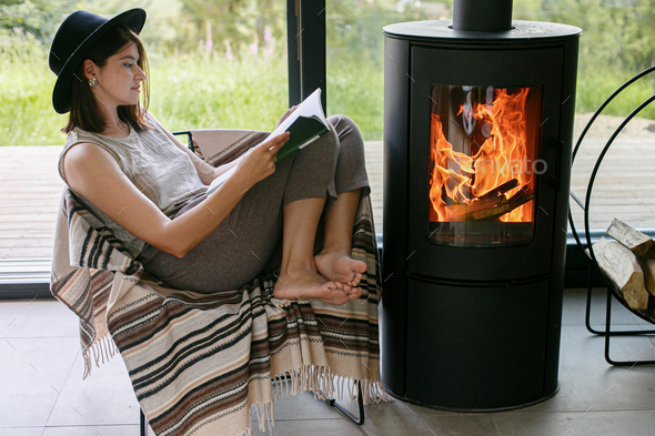 Beautiful stylish woman reading book on chair at warm burning fireplace, cozy vacation. Leisure time