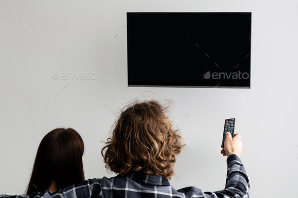 Couple Pointing Remote Control On Empty TV Screen Indoor, Back-View