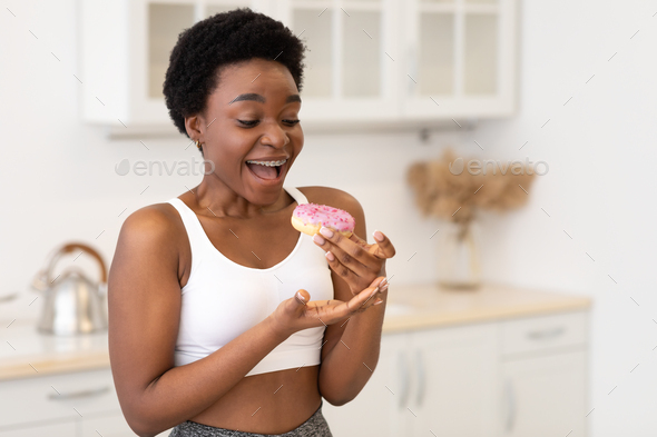 Excited Black Fitness Lady Eating Donut Standing In Modern Kitchen