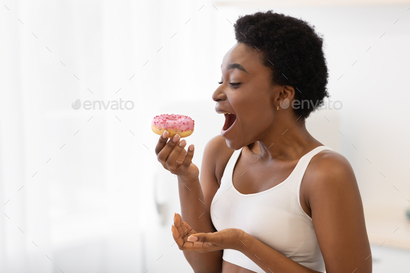 Excited Black Woman Eating Donut Having Cheat Meal Indoor, Side-View
