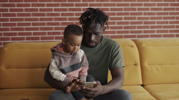 African American Father Sitting with Son on Sofa Using Smartphone alt