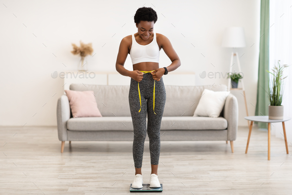 Joyful Black Female Measuring Waist Standing On Scales At Home