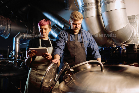 Young woman with digital tablet standing next to male colleague opening lid of huge steel cistern