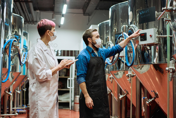 Young female brewery expert and male worker in masks testing new beer production equipment