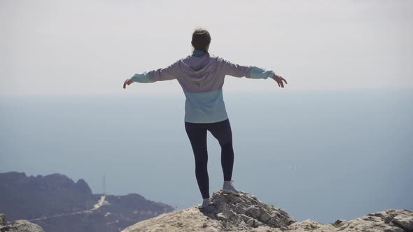 A Girl Stands on the Edge of a Cliff Against the Backdrop of the Sea and High Rocks alt