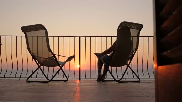 Young Woman Sits in a Sun Lounger on the Balcony with a Beautiful View of the Sea and Mountains alt