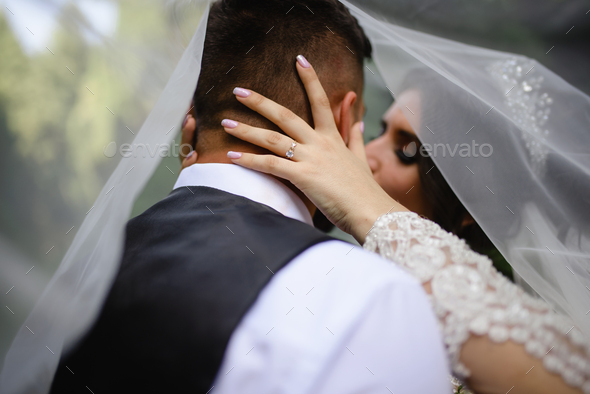 Close-up portrait of bride and groom under a veil. The bride kisses the groom.