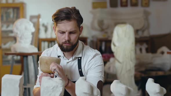 Bearded Stonemason Working in Studio with Mallet, Chisel and Limestone's Billets alt