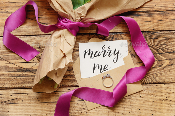 Bouquet in craft paper with a MARRY ME card and proposal ring on wooden table