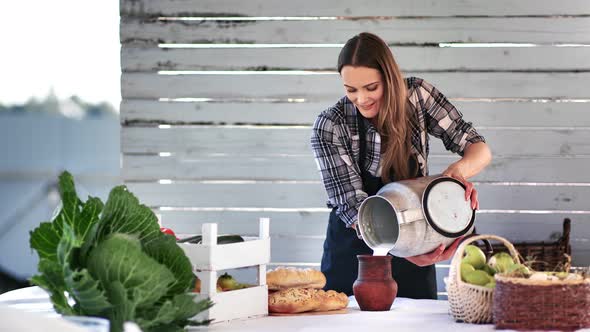 Smiling Farmer Female Pouring Homemade Milk From Metal Flask to Ceramic Mug alt