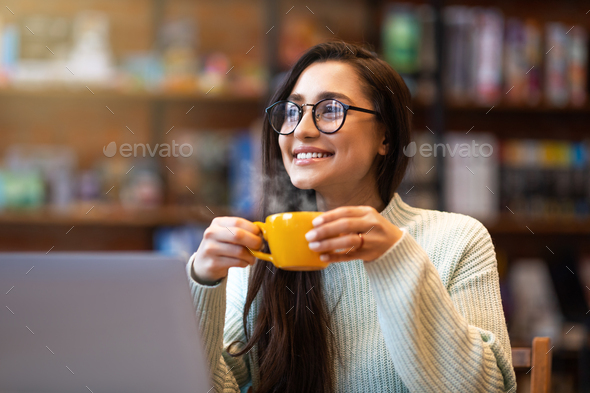 Benefits of remote work. Dreamy lady enjoying coffee during work on laptop in city cafe, smiling and - Stock Photo - Images
