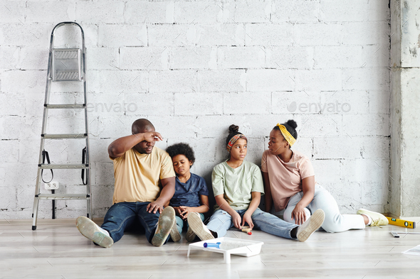 Tired African family sitting against wall on the floor of living-room after home renovation work