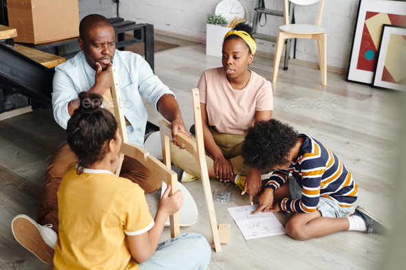 Puzzled African man looking at parts of chair while woman and boy pointing at assembling instruction - Stock Photo - Images