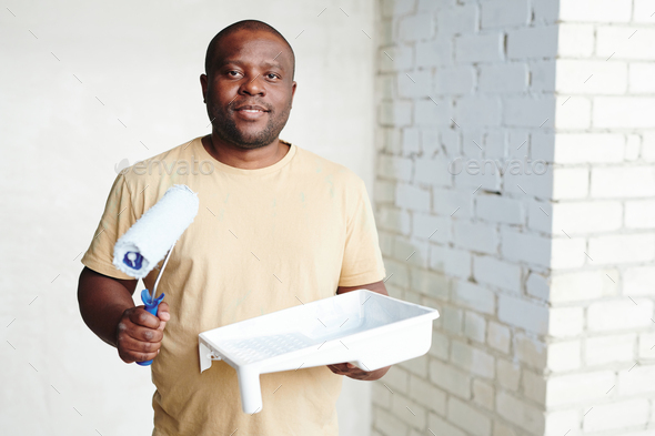 Happy African man with paintroller and white plastic container with paint standing against two walls - Stock Photo - Images