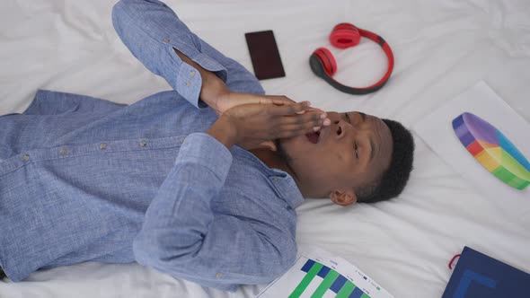 Top View of Exhausted African American Entrepreneur Lying on Bed with Graphs and Smartphone Touching alt