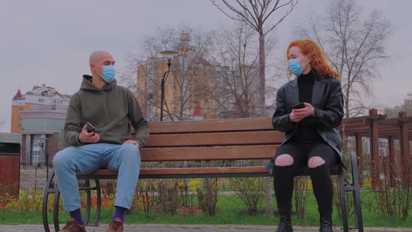 Man and Woman Sitting on Park Bench Take Off Their Protective Masks and Begin to Communicate alt