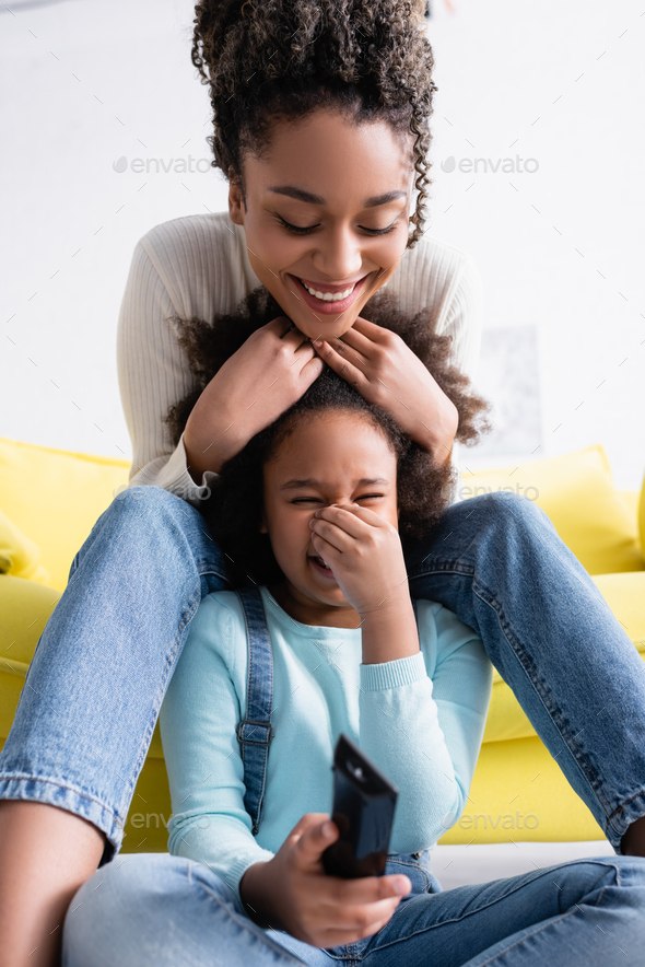 joyful african american woman touching head of laughing daughter watching comedy film