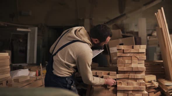 A Man in a Carpentry Workshop with a Tablet and a Pen Counts the Number of the Wood Blocks alt