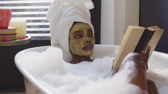 African american woman with towel and mask taking bath and reading book in bathroom alt