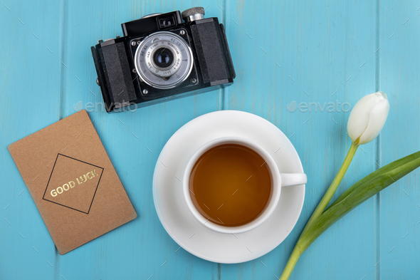top view of cup of tea on saucer and flower with photo camera and good luck card on blue background