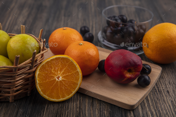 side view oranges with peach and cherry plum on a cutting board with plums in a basket on a wooden