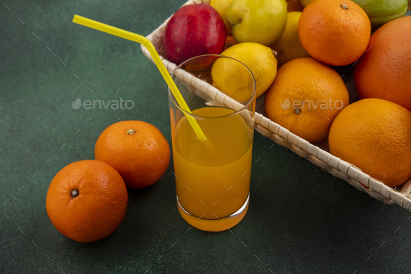 side view orange juice in a glass with oranges lemons and cherry plum in a basket on a green