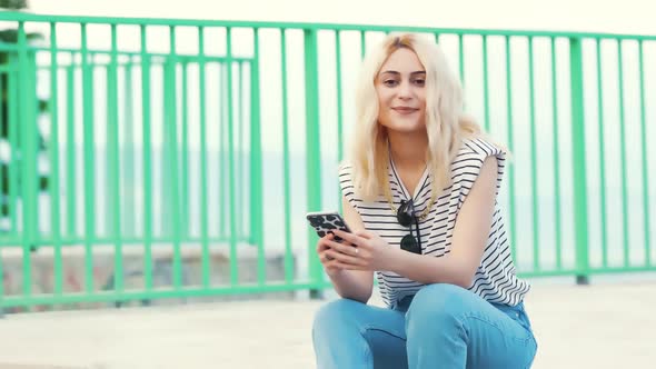 Pretty Blond Caucasian Girl Sitting Outside and Smiling Into the Camera Medium Full Shot Outdoor alt