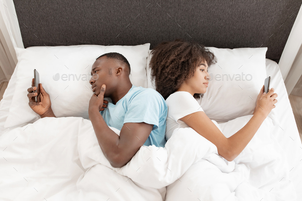 Awake using phone for chat, couple in quarrel, lying on bed back to back, using smartphones