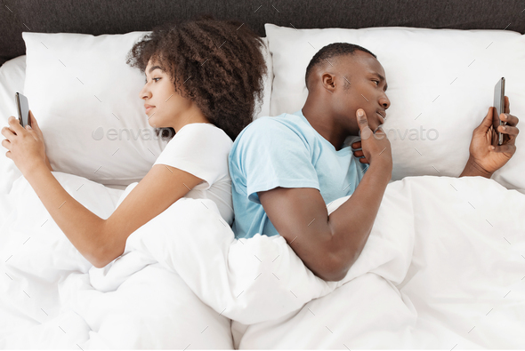 Couple using phones in bed, lying back to back, ignoring after quarrel