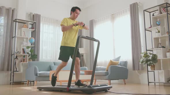 Asian Man Running On A Treadmill And Looking Smart Watch At Home alt