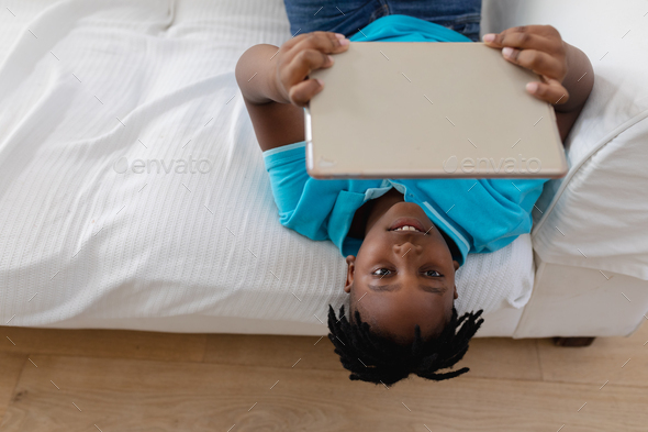 Smiling african american boy with short dreadlocks lying his back on couch using digital tablet