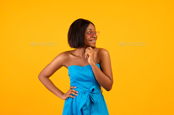 Pretty black lady in blue dress and sunglasses touching her chin and  looking aside on orange studio Stock Photo by Prostock-studio