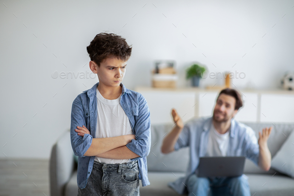 Furious father shouting at offended son while sitting with laptop computer on sofa at home