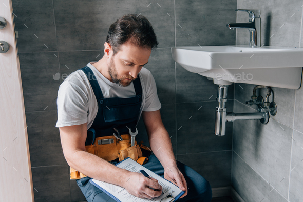 adult male plumber with toolbelt writing in clipboard near broken sink in bathroom