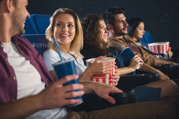 cheerful multiethnic friends with popcorn watching film together in movie theater