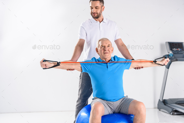 rehabilitation therapist assisting senior man exercising with rubber tape on fitness ball on grey