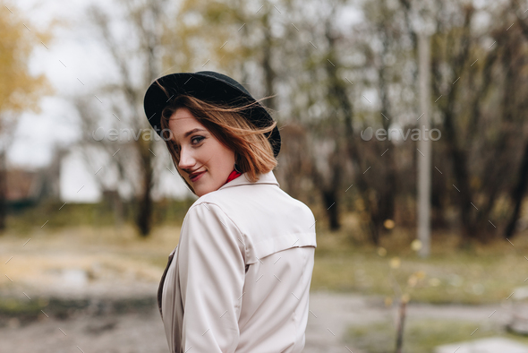 young smiling woman in trench coat and hat walking by autumn park and turning back