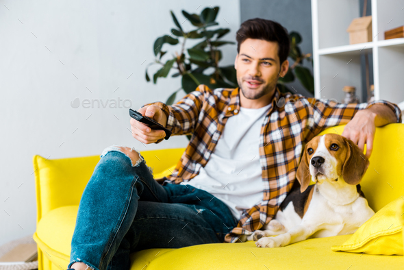 smiling man with remote control watching tv and sitting on sofa with dog