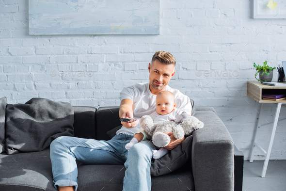 father sitting on couch with adorable baby daughter, holding remote controller and watching