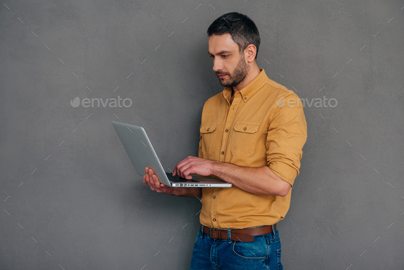 Confident IT expert. Confident mature man working on laptop while standing against grey background