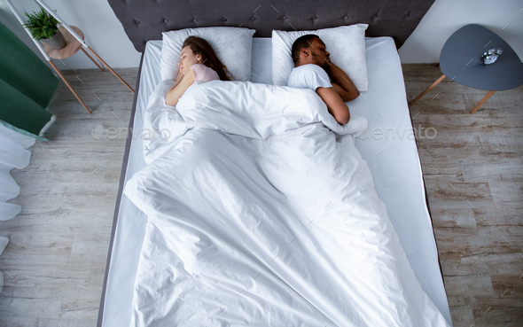 Millennial multiracial couple turning away from each other, sleeping in bed at home, top view