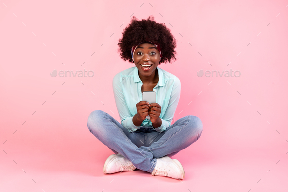 Excited black bushy lady using smartphone sitting over pink background