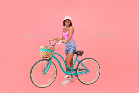 Full length of happy black lady in summer outfit standing with classic bicycle on pink studio