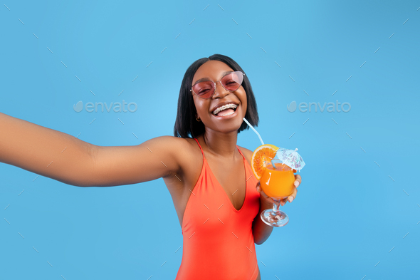 Happy black lady in bikini drinking tropical cocktail, taking selfie with smartphone on blue studio
