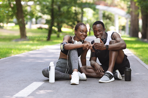 African Couple Using Fitness App On Smartphone Together Resting After Outdoor Training