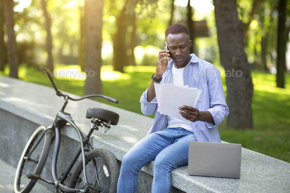 Worried black man with documents speaking on smartphone near his bike at city park
