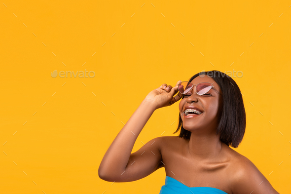 Portrait of happy black lady in dental braces touching sunglasses and smiling on orange background