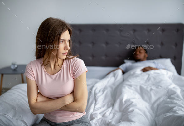 Young interracial couple having relationships crisis, depressed woman sitting on bed, her husband